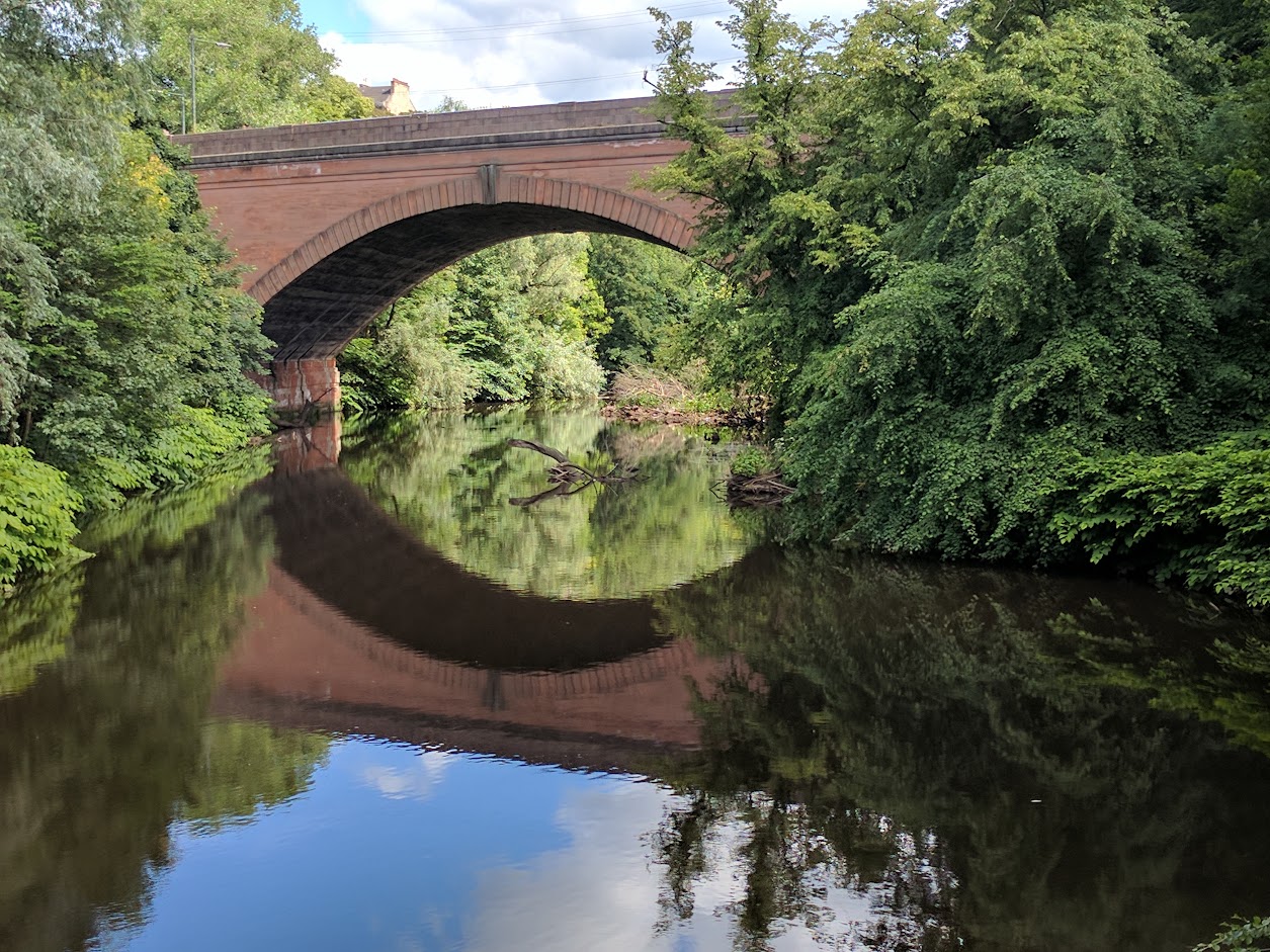 Stone bridge over stream surrounded by trees. Bridge is reflected in the still water.