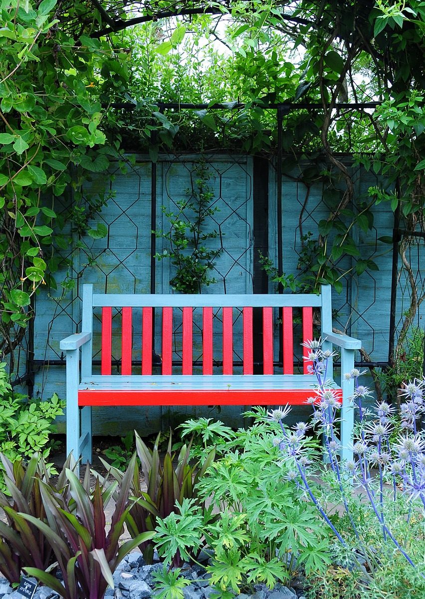 Red bench with blue arms and legs, sitting against a fence covered in climbing vines.